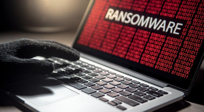 Mão com luva preta usando um laptop que exibe a palavra 'RANSOMWARE' em vermelho sobre um fundo com códigos binários, simbolizando uma ameaça de cibersegurança ativa.